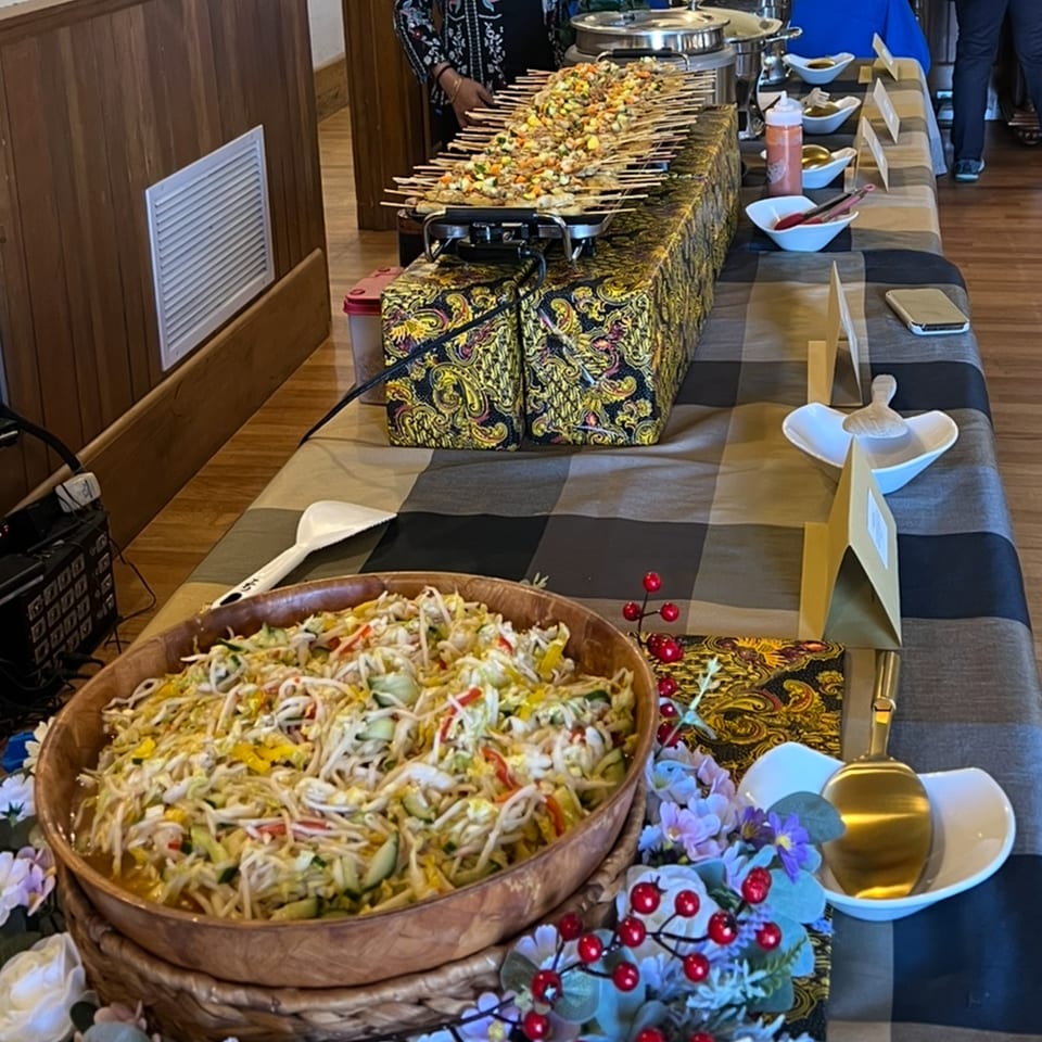 Indonesian buffet table with satay skewers, a large bowl of salad, and various serving dishes.