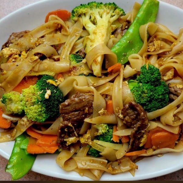 Indonesian stir-fried flat noodles with beef, broccoli, carrots, and snow peas in a white bowl.