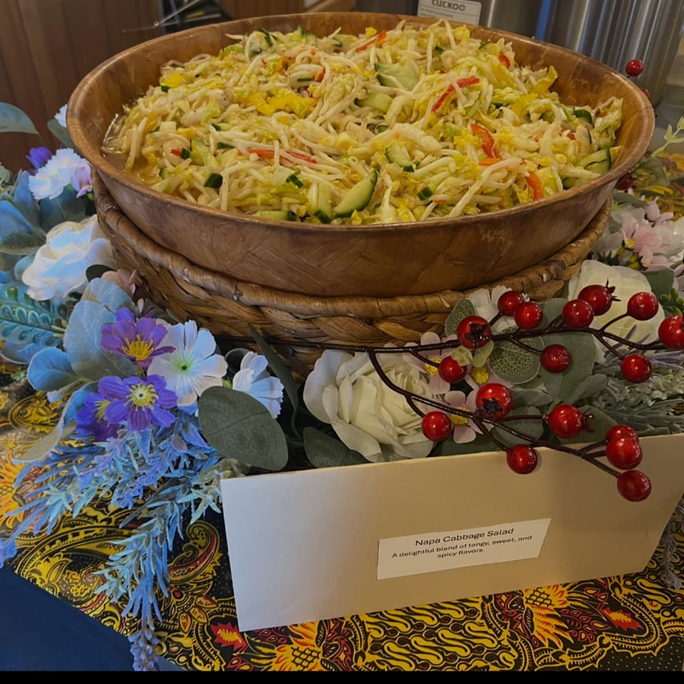 A large wooden bowl of Napa Cabbage Salad, garnished with flowers and berries, on a patterned tablecloth.