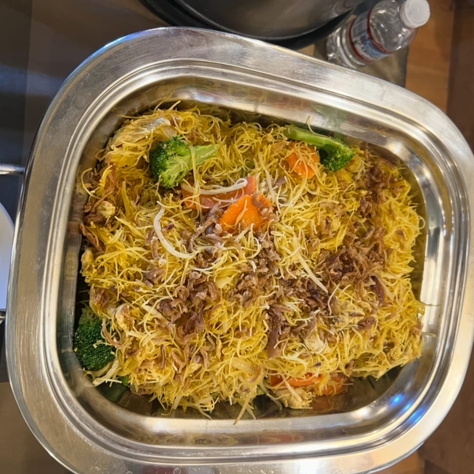 A metal serving dish filled with Indonesian noodles, broccoli, carrots, and fried shallots.