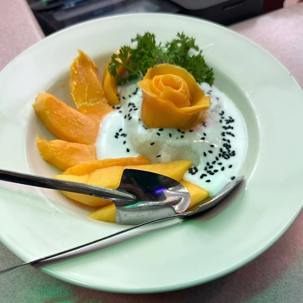 Mango sticky rice dessert with coconut milk, black sesame seeds, and parsley on a white plate.